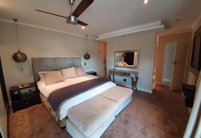 Cozy hotel bedroom with king bed, patterned headboard, hanging bedside lamps, bench seating, wall mirror, and neutral-toned decor