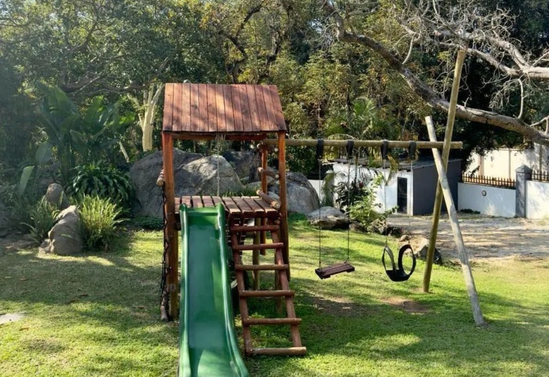 Outdoor wooden playground with green slide and swings set on lush green lawn surrounded by trees, ideal for family-friendly hotel guests