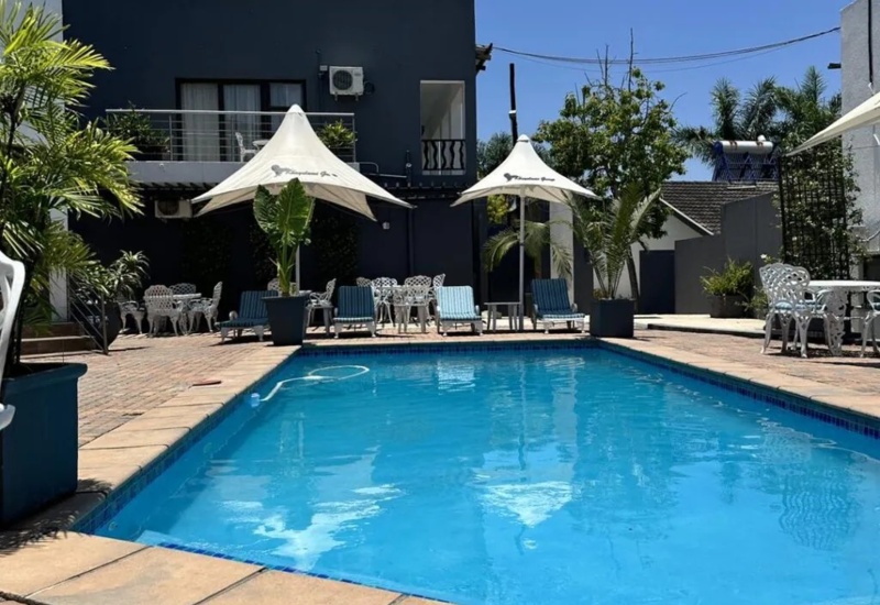 Outdoor swimming pool surrounded by lounge chairs with umbrellas, white patio furniture, and modern guest building under clear blue sky