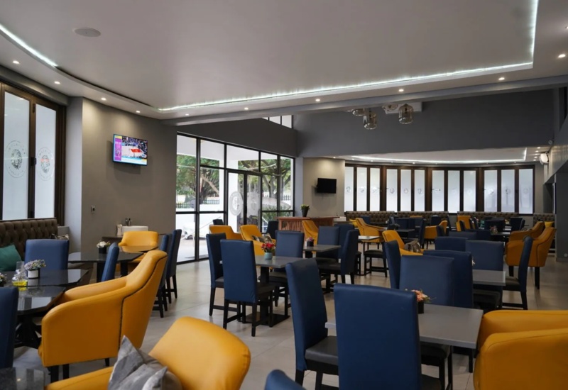 Modern hotel dining area with yellow and blue upholstered chairs, black tables, large windows, and ambient ceiling lighting