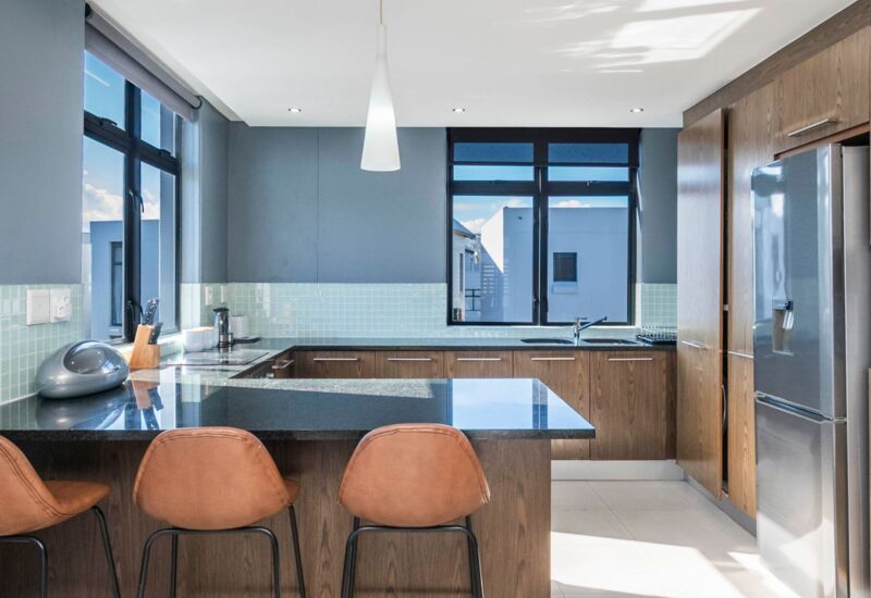 Modern kitchen with dark granite countertops, wood cabinets, three tan bar stools, large windows, and stainless steel refrigerator