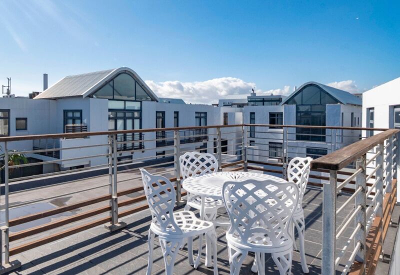 Sunlit rooftop terrace with white metal table and four chairs, surrounded by sleek railings and modern white buildings under blue sky