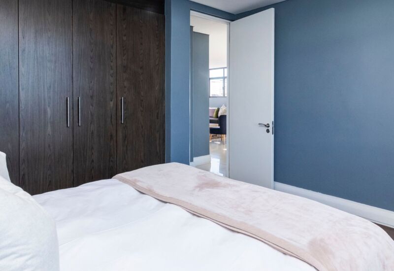 Modern hotel bedroom with navy blue walls, large dark wood wardrobe, white bedding, and soft beige throw blanket