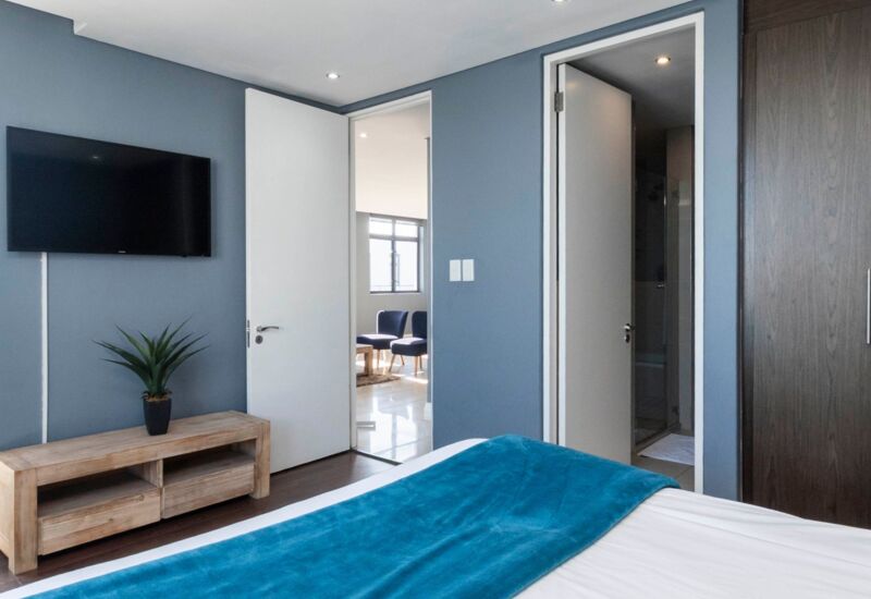 Modern hotel room with blue accent wall, wall-mounted TV, wooden console, blue throw on white bed, open doors revealing living area and bathroom