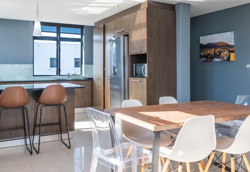 Bright modern kitchen and dining area with wooden cabinets, breakfast bar, clear and white chairs, and a cityscape photo on blue wall