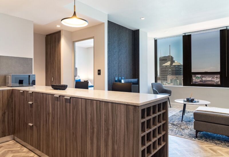 Modern hotel suite with wood kitchen island, marble countertop, dining area, and large window with cityscape view