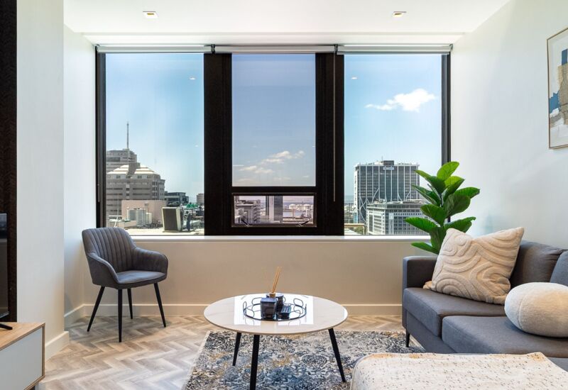 Modern hotel lounge with chevron wood flooring, large window showing city skyline, gray seating, patterned rug, and indoor plant