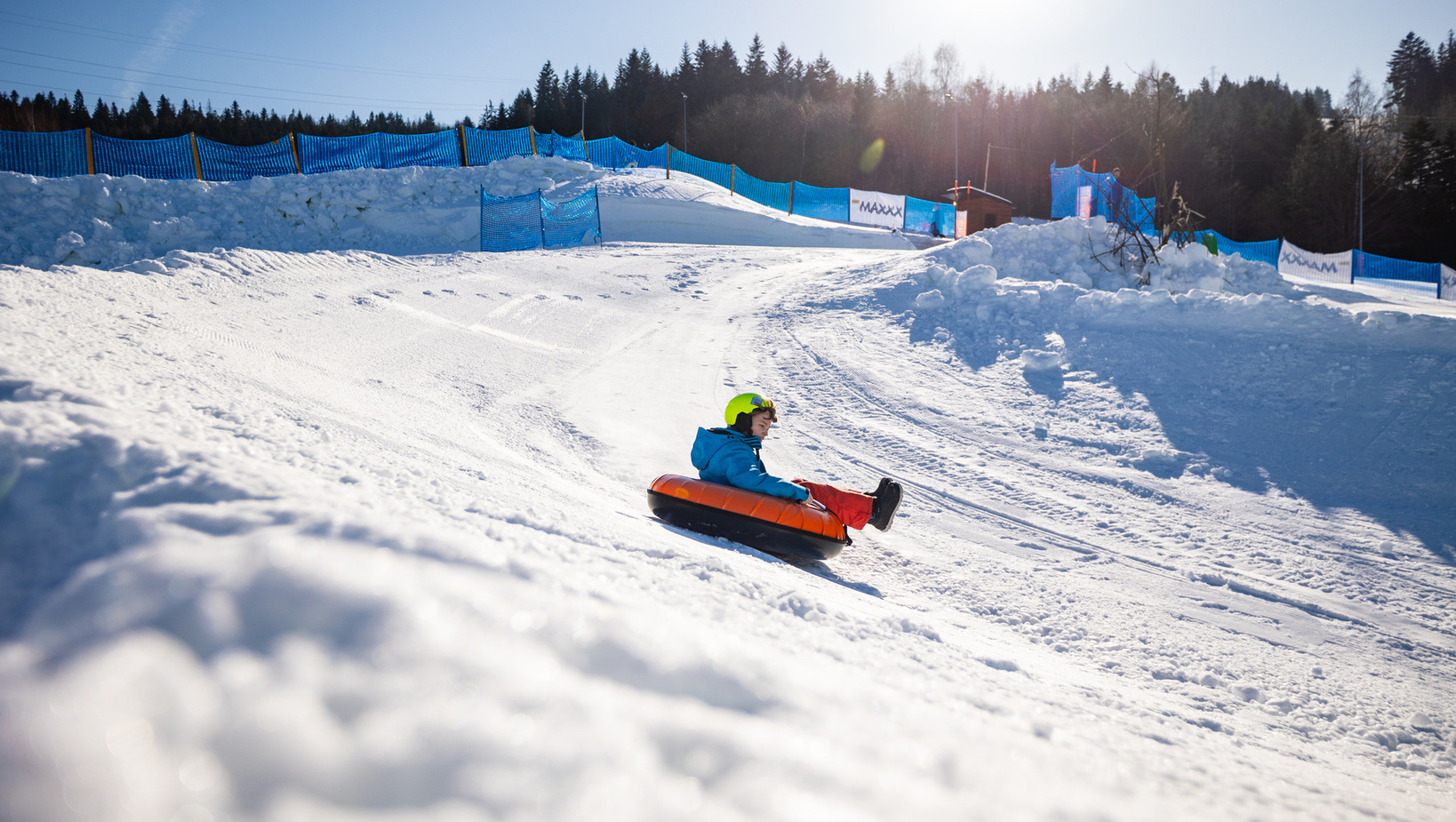 Child wearing helmet tubing down a snowy ski slope with blue safety nets and forest in the background on a sunny day.