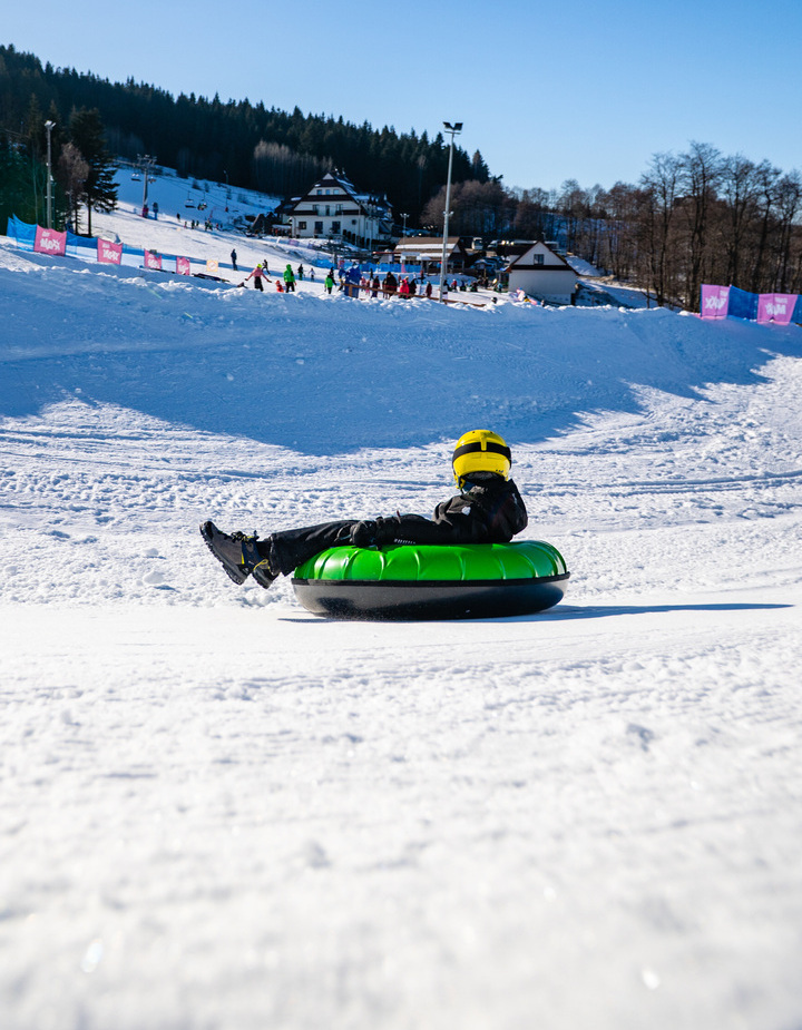 Guest wearing a yellow helmet sliding on a green snow tube down a snowy slope at a ski resort with lodges and ski lifts.
