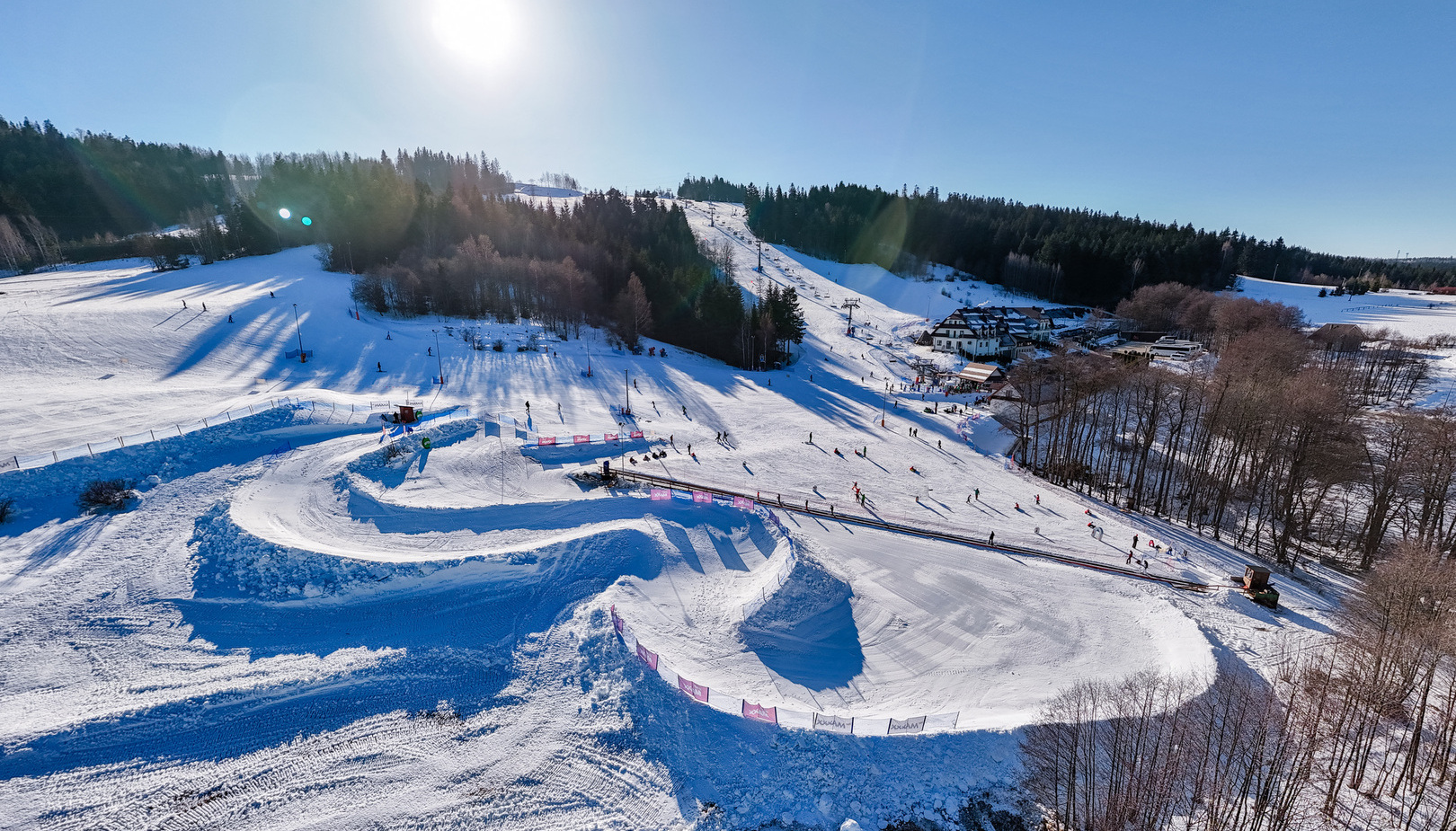 Ski resort with snowy slopes, chairlift, and sunlit forest in background, guests skiing and enjoying winter activities.