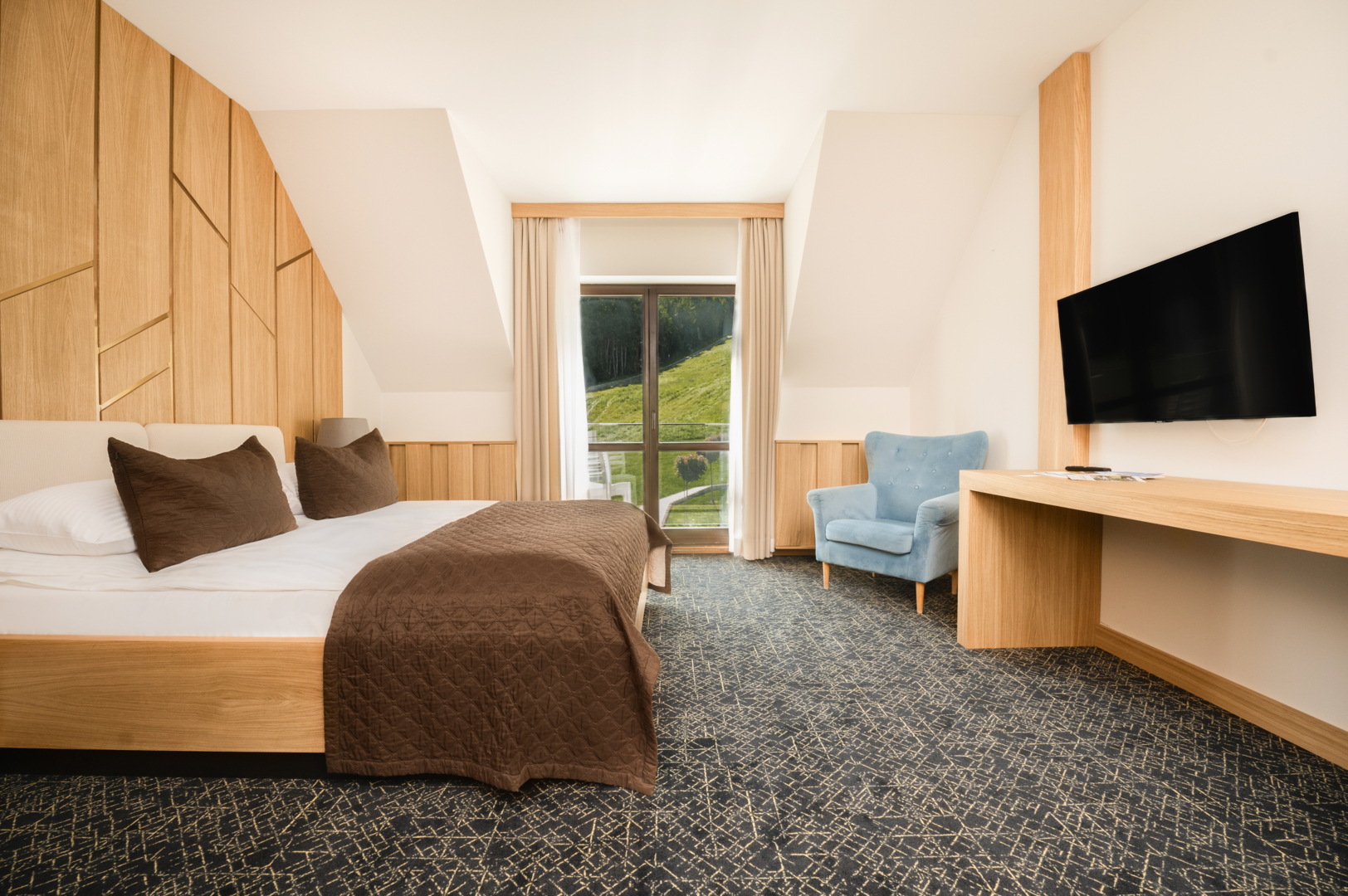 Nowoczesny pokój hotelowy z podwójnym łóżkiem, drewnianą ścianą, niebieskim fotelem i widokiem na zielony wzgórek przez okno.