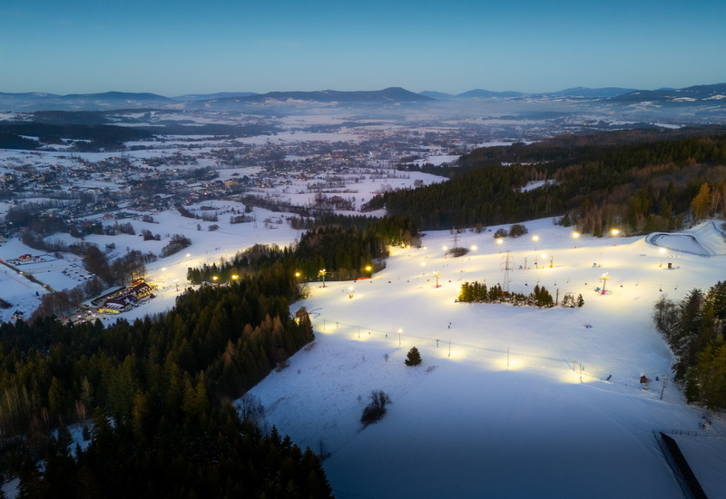 Illuminated ski slope on a snowy mountain with chairlifts and forest surroundings at dusk.