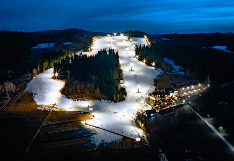 Ski resort with illuminated slopes and lifts, hotel building and parking surrounded by forest at dusk.