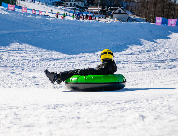 Guest wearing a yellow helmet slides down a snowy mountain slope on a green snow tube near the hotel.