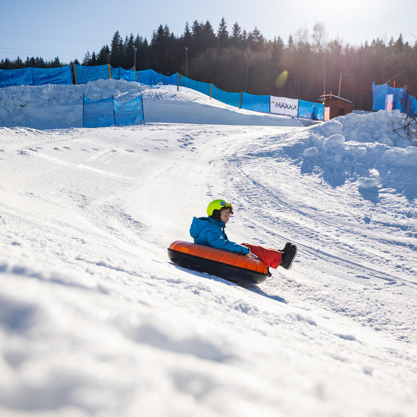 Dziecko z kaskiem zjeżdża na pontonie po zaśnieżonej trasie do snowtubingu z ochronnymi siatkami i widokiem lasu.