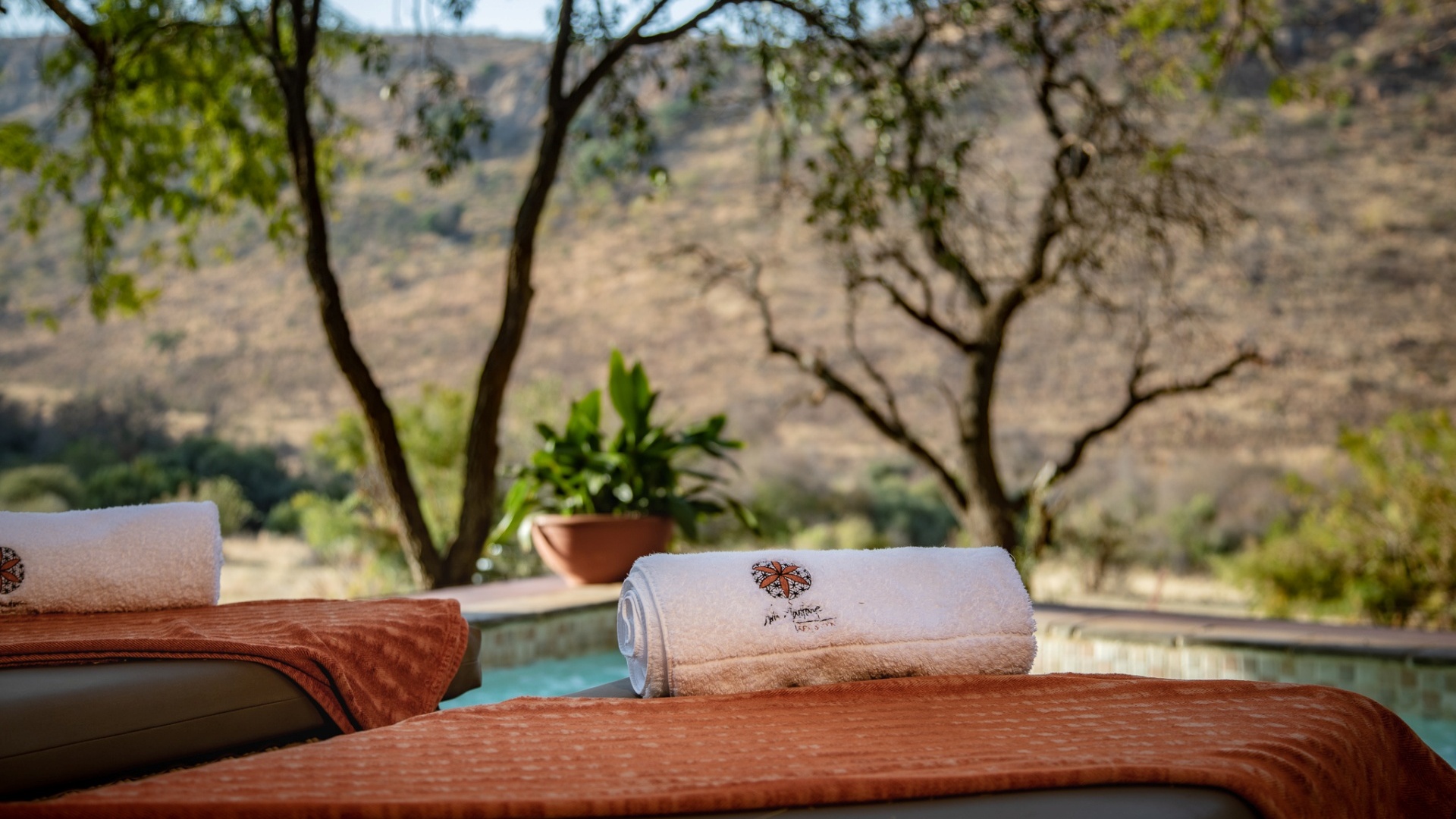 池畔躺椅配卷起的水疗毛巾和锈橙色毯，树荫下可眺望阳光照射的山坡与宁静的室外泳池。