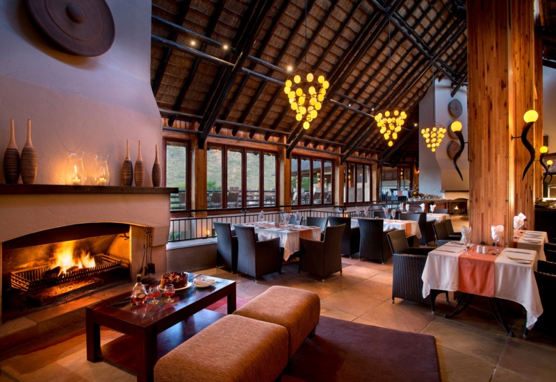 温馨茅草屋顶酒店餐厅，布置桌椅、藤编座椅、暖色球形吊灯、壁炉及可眺望山丘的露台。