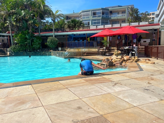 La Côte d’Azur outdoor pool with turquoise water, rock waterfall and Aqua Bar beneath red umbrellas; guest dipping hand.