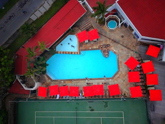 Aerial hotel pool deck with bright turquoise pool, hot tub, line of red umbrellas and loungers, adjacent tennis court