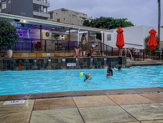 Hotel outdoor pool at dusk with children playing with a beach ball, elevated poolside bar lit purple, red umbrellas and terrace seating.