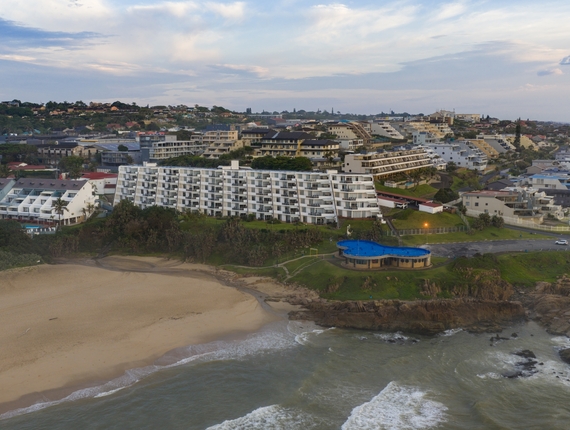 White beachfront hotel with balconies facing sandy beach, rocky shoreline and a blue-roofed pool pavilion on a grassy cliff.