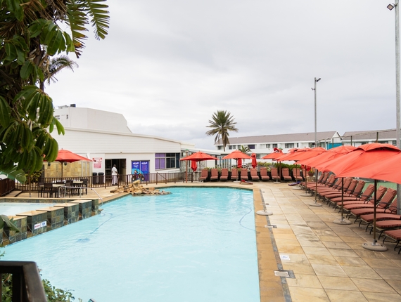 Outdoor La Côte d’Azur pool with turquoise water, rock waterfall, rows of red umbrellas and loungers on tiled deck, palm tree and poolside seating.