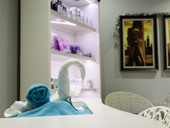 Spa reception with towel swan and rolled blue towels on white counter, illuminated product shelves and framed art.