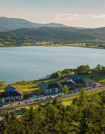Nowoczesny hotel nad jeziorem z widokiem na górskie krajobrazy, otoczony zielonym lasem i łąkami.