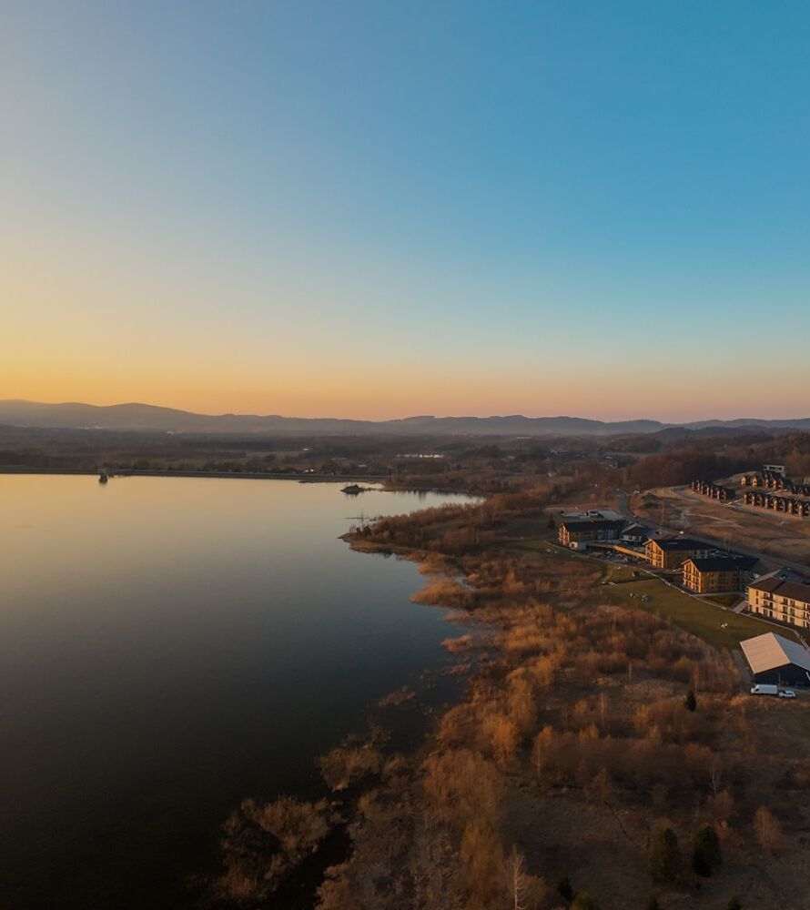 Widok na kompleks hotelowy nad jeziorem o zachodzie słońca z otaczającymi wzgórzami i lasem.