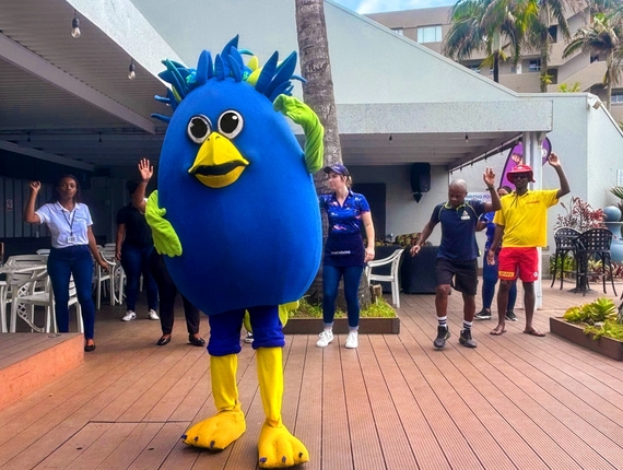 Blue bird resort mascot leading poolside activity with staff on wooden deck, palm trees and outdoor seating in background
