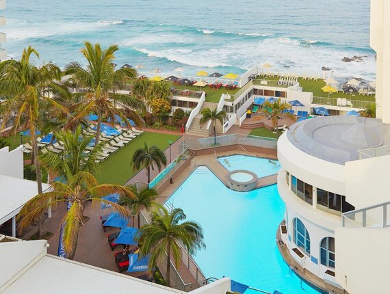 Oceanfront hotel pool and circular spa surrounded by palm trees, sun loungers, blue umbrellas and cliffside sundecks overlooking waves.