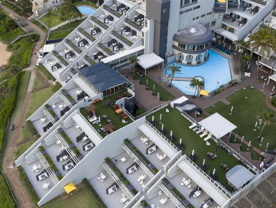 Tiered hotel terraces with private balconies by the beach, central pool with palms, lawn lounges and outdoor dining.