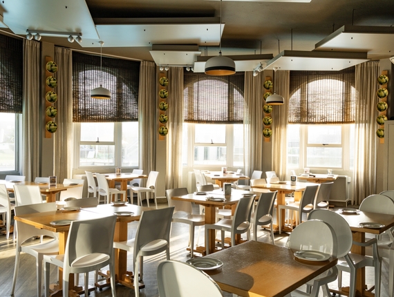Bright hotel dining room with wooden tables, white chairs, pendant lights and woven shades in warm morning light