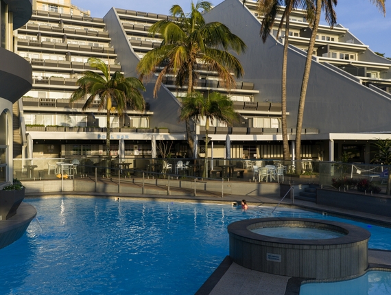 Resort pool with circular hot tub, palm trees and pool bar, modern tiered hotel façade, guest swimming near steps.