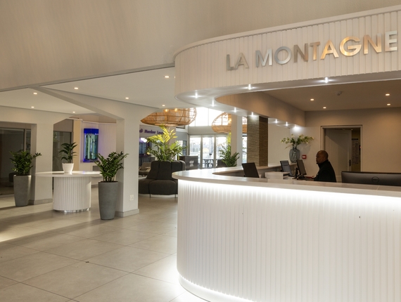 Modern La Montagne reception with illuminated curved white desk under 'LA MONTAGNE' sign, receptionist at desk, plants and lounge seating.