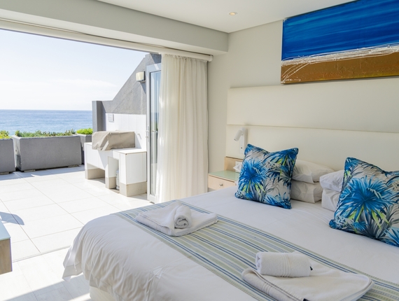 Ocean-view suite with king bed, blue tropical cushions and fresh towels, sliding doors open to sun terrace and sea.
