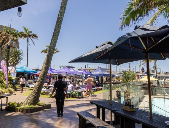 Busy La Montagne deck with shaded umbrellas, sun loungers, pool, palm trees and guests enjoying a market and swimming.