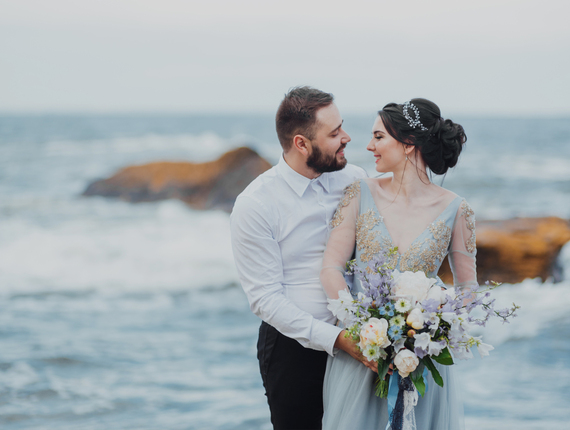 Bride and groom embracing on a rocky beachfront, bride in embroidered gown holding lush bouquet at a seaside wedding.