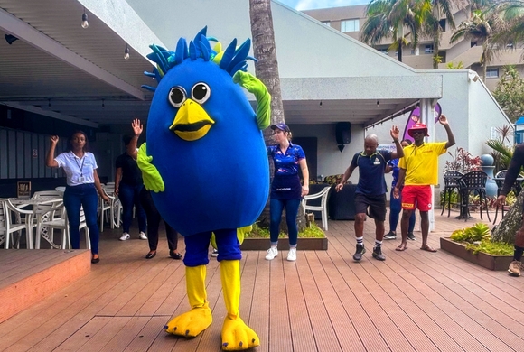 Blue bird resort mascot leading poolside activity with staff on wooden deck, palm trees and outdoor seating in background