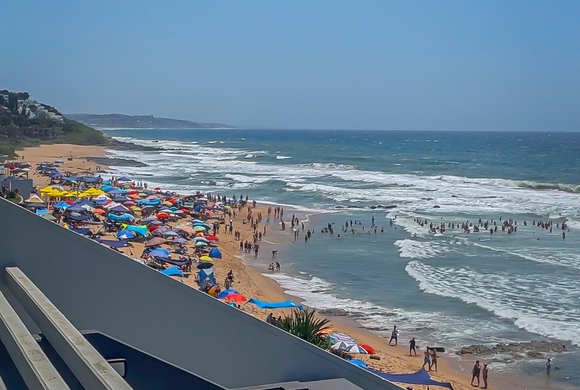 Balcony view over a lively sandy beach dotted with colorful umbrellas, swimmers in the surf, rolling waves and coastal homes.