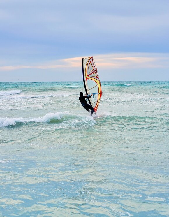 Windsurfer carving turquoise waves near shore under a soft pastel sky — active beachfront water-sports scene