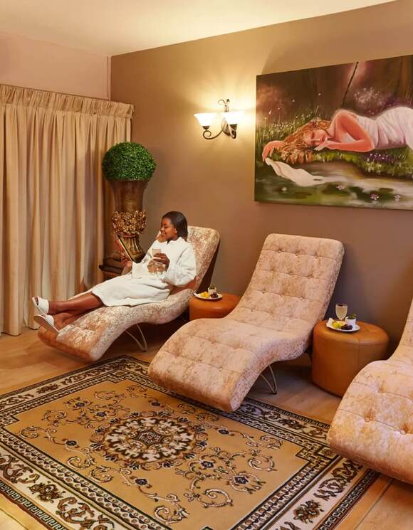 Spa relaxation lounge with three chaise lounges, soft lighting, decorative rug, refreshments; guest in robe sipping drink.