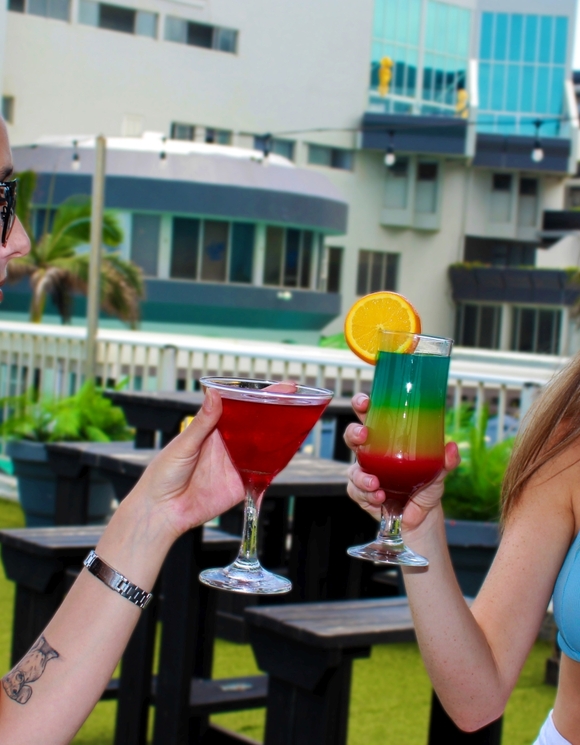 Two women in swimwear toasting colorful cocktails on a hotel's rooftop terrace with lounge seating, palm trees and modern beachfront buildings.