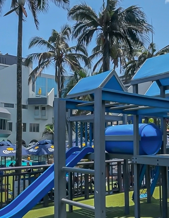 Resort poolside playground with blue slides and tunnel, palm trees, Corona-shaded lounge umbrellas and modern hotel building