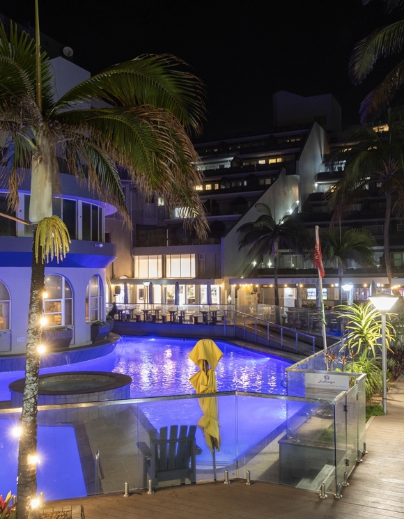 Nighttime La Montagne pool with blue LED-lit water, palm trees, glass balustrade, poolside deck chairs and lit restaurant terrace.