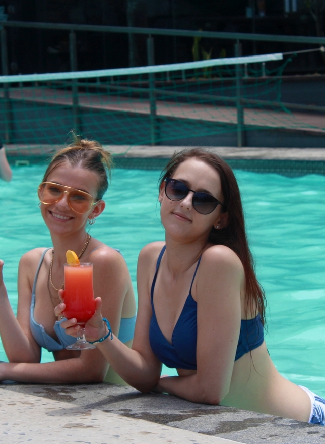 Two women in bikinis poolside at a hotel, holding colorful cocktails with orange garnish, wearing sunglasses.