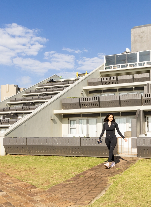 Stepped coastal hotel with private balconies, lawn and brick path; guest holding shoes walking from a ground terrace.