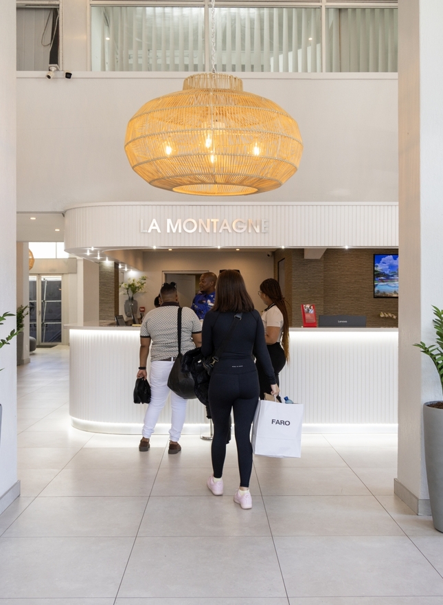 Bright modern lobby with reception desk 'LA MONTAGNE', guests checking in, woven pendant lights and tall potted plants.