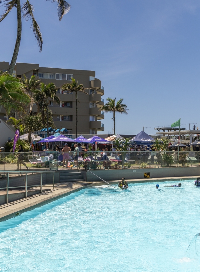 Sunny La Montagne pool with swimmers, glass railing, palm trees and lively poolside bars shaded by branded umbrellas.