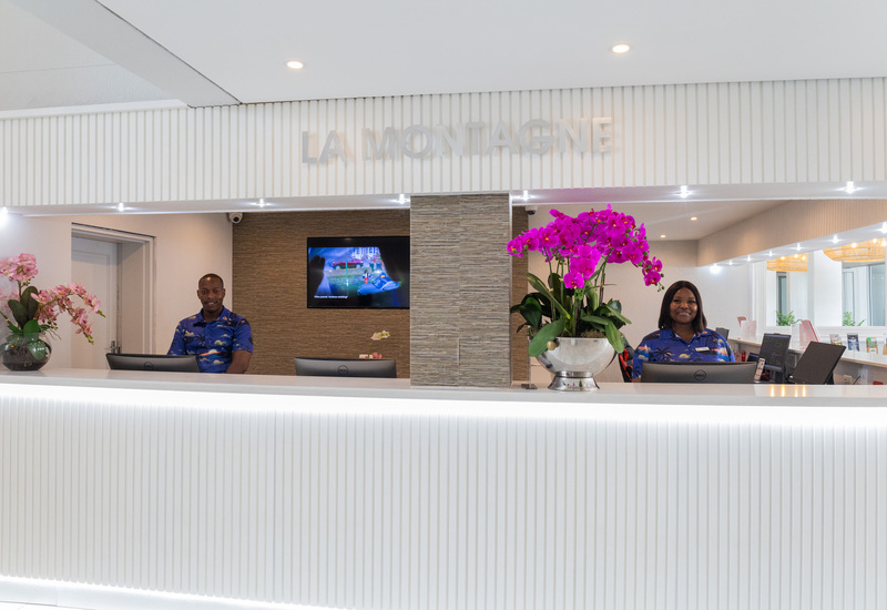 Bright modern hotel reception with white ribbed desk, vibrant purple orchids and two smiling staff at check-in under 'LA MONTAGNE' sign